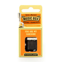 Load image into Gallery viewer, This image displays the bright orange and yellow cardboard packaging for a Kikkerland hand-crank music box, standing vertically against a plain white background. A silver metal crankshaft handle extends upward from the top left corner, while a small rectangular window on the front reveals the inner mechanical music parts directly beneath the printed song title, "YOU ARE MY SUNSHINE."