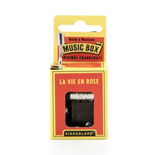 Load image into Gallery viewer, This image displays the dark red and yellow cardboard packaging for a Kikkerland hand-crank music box, standing vertically against a plain white background. A silver metal crankshaft handle extends upward from the top left corner, while a small rectangular window on the front reveals the inner mechanical music parts directly beneath the printed song title, "LA VIE EN ROSE."