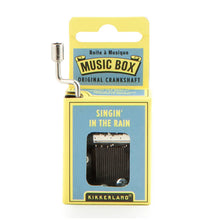Load image into Gallery viewer, This image displays the light blue and yellow cardboard packaging for a Kikkerland hand-crank music box, standing vertically against a plain white background. A silver metal crankshaft handle extends upward from the top left corner, while a small rectangular window on the front reveals the inner mechanical music parts directly beneath the printed song title, "SINGIN' IN THE RAIN."
