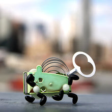 Load image into Gallery viewer, This image features a whimsical, mechanical wind-up toy with a sleek light green metal body standing on a flat gray surface against a blurry outdoor backdrop. The insect-like gadget showcases a large silver butterfly winding key extending from its rear, arched metal ribbon springs along its spine, and several jointed black legs positioned as if it is in mid-stride.