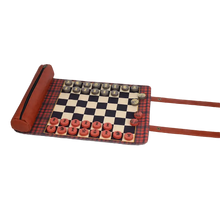 Load image into Gallery viewer, An unrolled plaid fabric chess and checkers board featuring flat red and gray game pieces arranged on the board alongside an attached zippered leather pouch and brown snap straps.