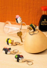 Load image into Gallery viewer, This image features five realistic cat-sushi keychains playfully staged on a light wooden table next to a Kikkoman soy sauce bottle, a clear glass cup, and a smooth wooden dome. The small figurines depict detailed cats perched on textured rice blocks like sushi toppings, with one keychain holding an actual brass door key that hangs over the side of the glass.