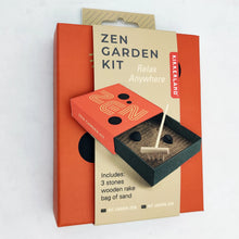 Load image into Gallery viewer, This retail box for a Kikkerland "Zen Garden Kit" features a prominent orange background wrapped by a central, beige paper sleeve. The packaging displays an illustration of a tiny, dark green matchbox-style tray filled with sand, a miniature wooden rake, and two smooth black stones, positioned below the words "Zen Garden Kit: Relax Anywhere."