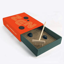 Load image into Gallery viewer, This image displays the Kikkerland "Zen Garden Kit" out of its main packaging, showing the tiny matchbox-style tray partially slid open against a plain white background. The slide-out tray is a dark forest green and filled with fine sand, containing two small, smooth black stones and a miniature light-wood rake, while the orange outer sleeve features the word "ZEN" in a stylized, multi-lined font.
