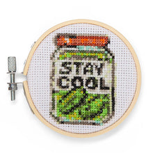 Load image into Gallery viewer, This image displays a completed cross-stitch project of a glass jar filled with green pickles isolated against a clean white background. Framed within a small, circular wooden embroidery hoop, the design features a red lid and a white center label with the words "STAY COOL" stitched in black block lettering.