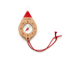 Load image into Gallery viewer, This image shows the unpackaged "Huckleberry Compass" resting flat against a clean, white background. The leaf-shaped wooden compass features carved directional initials around its clear center dial, a solid bright red painted tip at the top, and a matching red cord lanyard looped through the bottom.