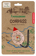 Load image into Gallery viewer, This image shows the retail packaging for a children's "Huckleberry Compass," featuring a brown cardboard backing illustrated with rustic mountain peaks and a winding blue river trail. Attached to the center is the actual compass, which is housed in a light-colored, leaf-shaped wooden casing with a bright red tip at the top and a black elastic lanyard at the bottom.