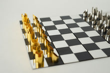 Load image into Gallery viewer, A close-up side view of a modern chess set featuring metallic gold and silver cylindrical pieces set up on a black-and-silver checkered board.