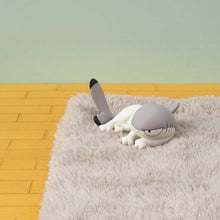 Load image into Gallery viewer, A single grey-and-white Sharkitty figurine lounging on a grey mini rug. It rests on its belly with half-lidded eyes and its tail pointing straight up in the air against a yellow wooden floor and soft green wall.