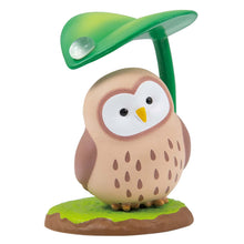 Load image into Gallery viewer, A round, light brown owl figurine with speckled chest feathers and a heart-shaped white face, standing on a grassy base against a plain white background. The owl perches under a green leaf that curves over its head like a small umbrella, holding a single clear raindrop on its surface.