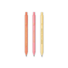 Load image into Gallery viewer, three soft-touch gel pens are lined up vertically side-by-side. The pens come in a warm pastel palette of coral, pink, and pale cream, with each barrel featuring a playful phrase cleanly printed lengthwise in a subtle, metallic gold font.