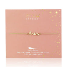 Load image into Gallery viewer, This image shows a gold "Aries" script bracelet mounted flat against a warm, peach-colored square card decorated with metallic gold stars and dots. The minimalist bracelet features a delicate cursive nameplate charm dipped in 14k gold, threaded onto a slim, adjustable tan cord.