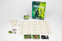 Load image into Gallery viewer, a game setup for Codenames Duet, displayed featuring twenty-five white word cards arranged in a flat five-by-five grid on a light background, with the green game box standing upright behind them. Placed around the grid are two thick stacks of illustrated green agent tiles, extra card decks, small tokens, a dark assassin card, and a small keycard mounted upright on a stand.