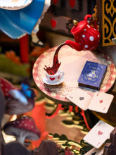 Load image into Gallery viewer, A close-up view depicts a small wooden round table setting with playing cards, a book, and a red teapot pouring tea directly into a heart-decorated cup.