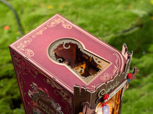 Load image into Gallery viewer, An overhead angled view shows the top of the dark red outer box casing, which features intricate gold foil flourishes and a keyhole-shaped viewing window looking down inside the book nook.