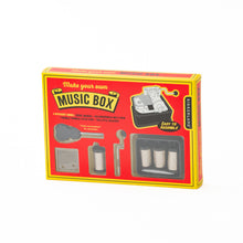 Load image into Gallery viewer, This image displays a rectangular, red and yellow cardboard box for a "Make your own Music Box" kit, sitting at an angle against a white background. The front of the box features several small plastic-covered cutout windows that neatly display the individual unassembled components inside, including a hand-crank handle, musical combs, a screwdriver, and different pin-textured cylinders.