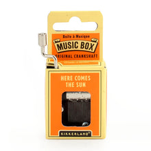 Load image into Gallery viewer, This image shows an identical small, hand-cranked music box package, but this version features a bright orange and pale yellow cardboard design. A silver metal crank sticks out of the top left, and the text on the front orange panel reads "HERE COMES THE SUN" right above the clear window displaying the metal musical comb.
