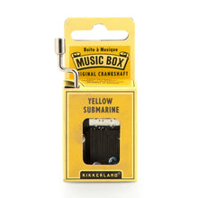 Load image into Gallery viewer, This image displays the entirely yellow cardboard packaging for a Kikkerland hand-crank music box, positioned vertically against a plain white background. A silver metal crankshaft handle extends upward from the top left corner, while a small rectangular window on the front reveals the inner musical mechanism directly beneath the printed song title, "YELLOW SUBMARINE."
