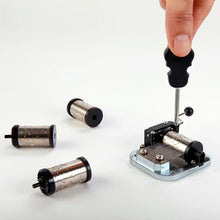 Load image into Gallery viewer, This image shows a person's hand using a black, violin-shaped screwdriver to assemble an exposed metal music box mechanism on a clean white surface. To the left of the main mechanism, three separate, loose silver cylinders with tiny textured metal pins lie scattered on the table.
