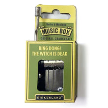 Load image into Gallery viewer, This image displays the olive green and yellow cardboard packaging for a Kikkerland "Music Box" with a crank handle sticking out from the top left corner. The front features a central cutout window that shows the silver metal musical comb and pinned cylinder inside, right below the printed song title: "DING DONG! THE WITCH IS DEAD."
