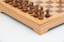 Load image into Gallery viewer, A close-up view showing dark wooden chess pieces set along the corner of a handcrafted wooden board with carved trim and feet.
