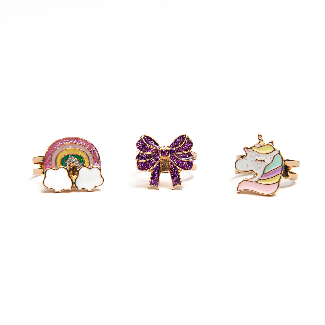 Set against a stark white background, there are three gold-colored novelty rings for children arranged neatly in a row. From left to right, the ring faces include a pink-glittered rainbow resting between two white clouds, a sparkling purple bow, and a unicorn head with a soft pastel mane.