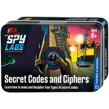 Load image into Gallery viewer, This image features the front of a small, rectangular storage tin for a children's activity set titled "Secret Codes and Ciphers," intended for ages 8 and up under the "Spy Labs Incorporated" brand. The box art displays a vibrant, comic-style illustration from a detective's point of view, showing a hand holding a circular black-and-red cipher wheel with alphabet letters against a city street backdrop at sunset.
