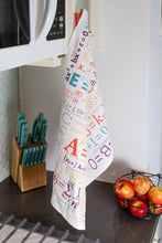 Load image into Gallery viewer, A white kitchen towel printed with colorful mathematical and scientific equations hangs from a cabinet next to a knife block and a wire basket of apples.
