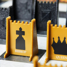 Load image into Gallery viewer, A close-up view of gold castle-turret-shaped game pieces featuring printed black silhouettes and names for the "BISHOP" and "QUEEN".
