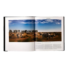 Load image into Gallery viewer, Open interior spread of a book showcasing a wide panoramic photograph of a large herd of rhinoceroses standing in a dry, grassy African landscape under a blue sky.
