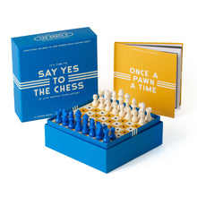 Load image into Gallery viewer, A blue "SAY YES TO THE CHESS" product box standing next to the yellow instruction book and a raised box-frame chessboard set with blue and cream peg chess pieces.
