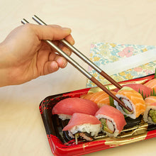 Load image into Gallery viewer, This image captures a hand utilizing a pair of modern chopsticks to pick up a piece of sushi from a red-and-black plastic tray filled with various rolls and nigiri. The chopsticks feature sleek, silver metallic upper handles seamlessly joined to natural brown wooden tips, set against a light wood-grained tabletop with a floral paper napkin nearby.
