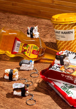 Load image into Gallery viewer, This image shows five cat-sushi keychains scattered on a wooden surface next to instant ramen items and a bottle of prickly ash oil. The detailed figurines feature different cat breeds posing as sushi toppings bound to a rice block, with whimsical additions like tiny dollar bills, chopsticks, or a small blue flower hat.

