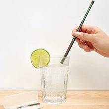 Load image into Gallery viewer, This image shows a person's hand holding the fully extended, iridescent rainbow-colored travel straw inside a clear glass of sparkling water garnished with a fresh lime wheel. The scene is set on a light wood tabletop, where the open, frosted plastic carrying case lies nearby with its small cleaning brush resting inside.
