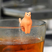 Load image into Gallery viewer, This close-up shot captures a tiny, solid-orange plastic cat figurine perched delicately on the condensation-covered rim of a clear glass mug. The miniature cat faces forward with tiny dotted black eyes and a nose, its body straddling the edge while holding a thin white tea bag string behind its tail in the warm, amber tea.
