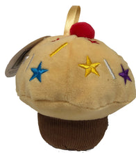 Load image into Gallery viewer, Set against a solid white background, a single plush vanilla-themed cupcake baby toy. The toy features a dark brown ribbed base mimicking a liner, a fluffy pale-yellow top decorated with multi-colored embroidered stars and sprinkles, and a small, fuzzy red pom-pom cherry stitched directly below a soft gold satin loop ribbon.
