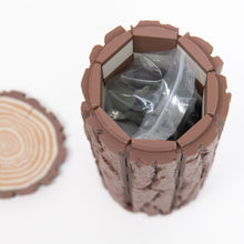 Load image into Gallery viewer, A hollow, tree-log-style container stands vertically with its wood-grain lid removed to reveal packaged game pieces inside.
