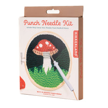 Load image into Gallery viewer, This retail box for a Kikkerland "Punch Needle Kit" features a nature theme and is shown from a slight angle, topped with a solid brown hanging tab. The white packaging prominently displays an illustration of a textured, completed project showcasing a large red and white spotted mushroom growing on green grass, all framed inside a six-inch wooden hoop.
