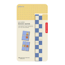 Load image into Gallery viewer, This image shows the retail packaging for Kikkerland's Inkerie "Pen Holder Book Band," displayed on a cream-colored cardboard backing with a yellow dotted section at the top. The product features a long, vertically arranged elastic band with a blue and off-white checkered pattern, positioned next to a couple of diagrams illustrating how to wrap the band around a notebook to hold pens.
