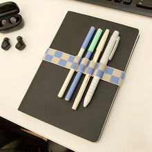 Load image into Gallery viewer, This image shows a closed, black hardcover notebook lying flat on a white desk with four pens secured to its front cover. The pens—colored light green, blue, cream, and grey—are held neatly in place by a blue and beige checkered elastic book band wrapped horizontally around the notebook's middle.
