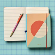 Load image into Gallery viewer, This image shows the open contents of the "Lined Mini Notebook &amp; Pen" set resting on a blue grid surface. It displays a slim, orange and purple color-blocked ballpoint pen with a gold tip and clip lying across an open notebook with lined pages, right next to a closed companion notebook that features a pastel peach and teal geometric design secured by a bright orange vertical elastic band.
