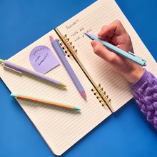 Load image into Gallery viewer, This image captures a person's hand in a purple sweater using one of the pens to write a to-do list in an open, gold spiral-bound notebook on a bright blue table. Several other pens from the set are scattered across the lined pages, which also feature a small lavender sticky note marked "URGENT" next to a handwritten list.
