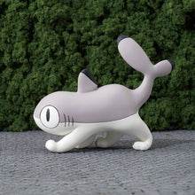 Load image into Gallery viewer, A single grey-and-white Sharkitty figurine prowling low on a grey paved surface, its tail held high in the air behind it. It is set against a lush green hedge in the background.
