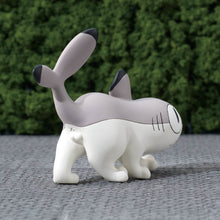 Load image into Gallery viewer, The rear three-quarter view of the grey-and-white Sharkitty figurine prowling forward on a grey surface, highlighting its raised tail fin and a tiny black "x" drawn beneath it to resemble a cats butt. A dense green hedge fills the background..
