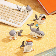 Load image into Gallery viewer, Five grey-and-white Sharkitty figurines resting across a textured yellow surface, with one lounging directly atop a white computer keyboard. They are surrounded by desk items including a bright orange notebook, a cardboard box, and a clear water bottle, creating a playful workspace setup.
