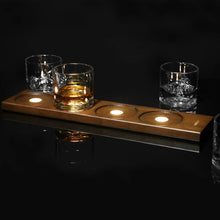 Load image into Gallery viewer, Set against a dark, reflective black background, a premium wooden whiskey flight board equipped with built-in, glowing circular lights inside each of its four glass holders. One clear glass filled with amber-colored liquor sits directly over a light on the board, while three other empty glasses, featuring unique molded mountain peaks inside their bases, rest nearby on the reflective surface.
