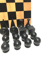 Load image into Gallery viewer, A close-up view of geometric black chess pieces—including spheres, cones, cubes, and scrolls—arranged in front of a wooden chessboard.

