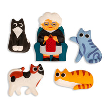 Load image into Gallery viewer, five colorful, cartoon-style magnets are featured arranged against a plain white background, centered around an elderly white-haired woman with glasses sitting in a blue armchair and knitting with red yarn. Surrounding her are four playful, stylized cats in various poses: a sitting black-and-white tuxedo cat, a stretching light-blue tabby, a walking calico, and a lounging orange striped cat.
