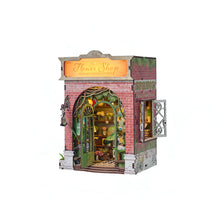 Load image into Gallery viewer, An angled front view shows a brick-façade miniature book nook depicting a flower shop with an illuminated sign board, an open green arched door, and a side window lattice.
