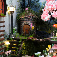 Load image into Gallery viewer, A detailed view features a glowing miniature streetlamp next to a small thatch-roofed roundhouse connected by a tiny wooden rope bridge surrounded by lush green moss.
